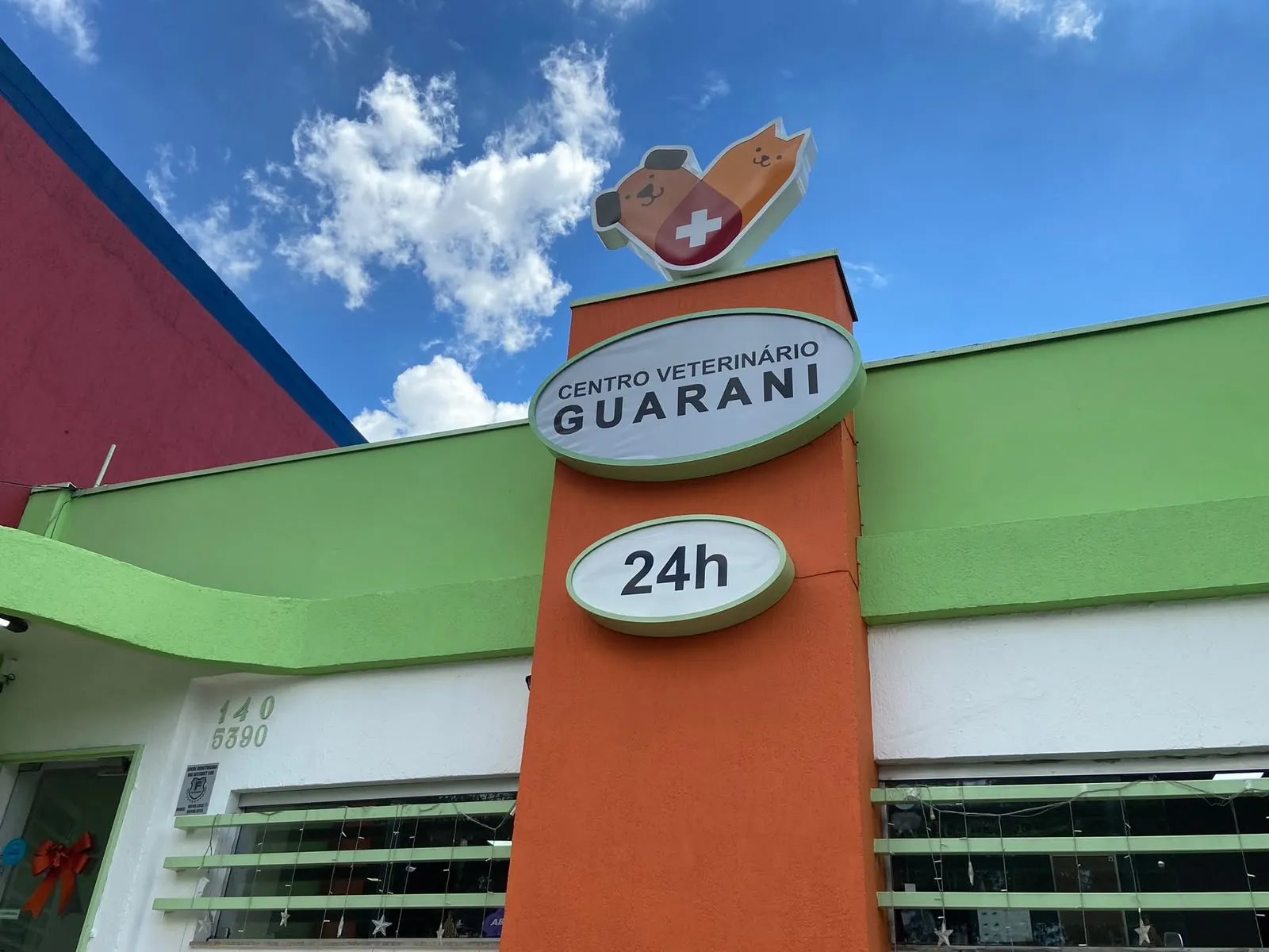 Totem do Centro Veterinário Guarani com a placa 24h, contra o céu azul, na fachada verde e branca da clínica