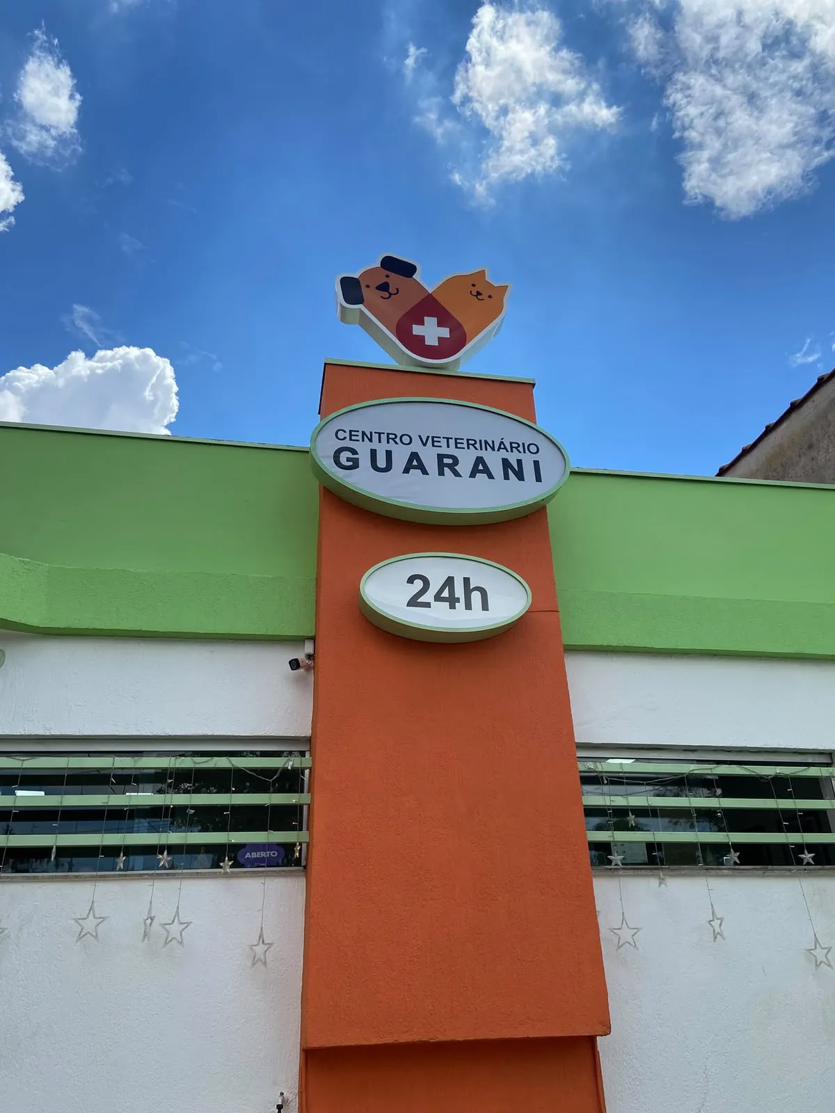 Totem laranja do Centro Veterinário Guarani visto de baixo, com o letreiro 24h, a fachada verde e o céu azul