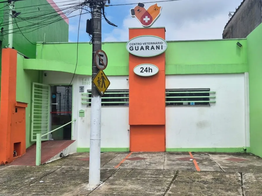 Fachada do Centro Veterinário Guarani vista da rua, com o totem laranja do letreiro 24h e a porta de vidro. Em frente à casa fica o recuo, com as 3 vagas marcadas no piso.
