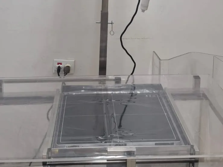 Sala de raio X da clínica: a mesa de exame, de acrílico transparente, com uma placa cinza no centro, a coluna de metal do aparelho e a tomada na parede branca