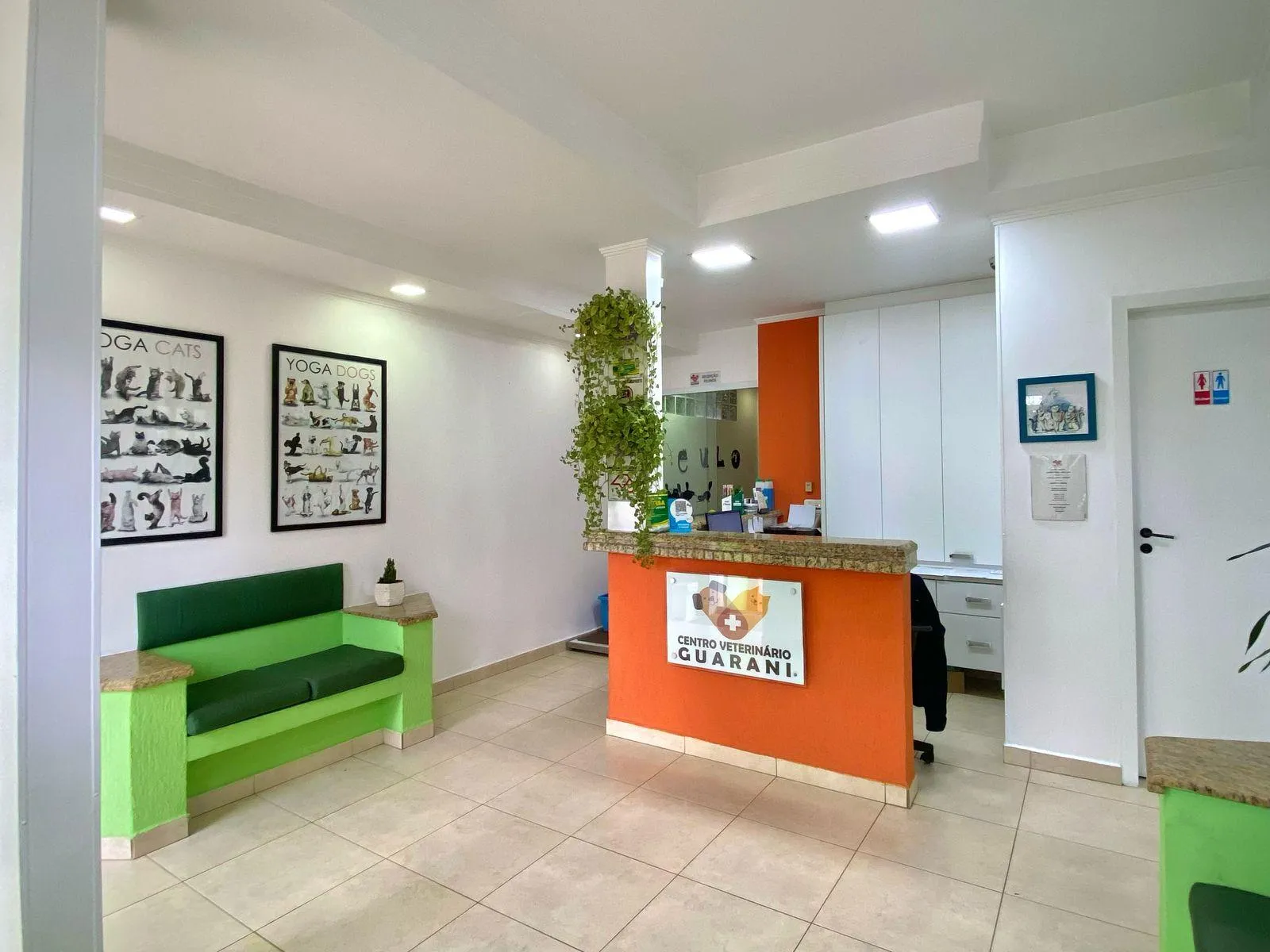 Recepção da clínica, com balcão laranja, bancos verdes e quadros de pets na parede